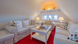 Una sala de estar acogedora con alfombra roja, sofás blancos con cojines de lunares, una mesa de centro de vidrio y dos lámparas.