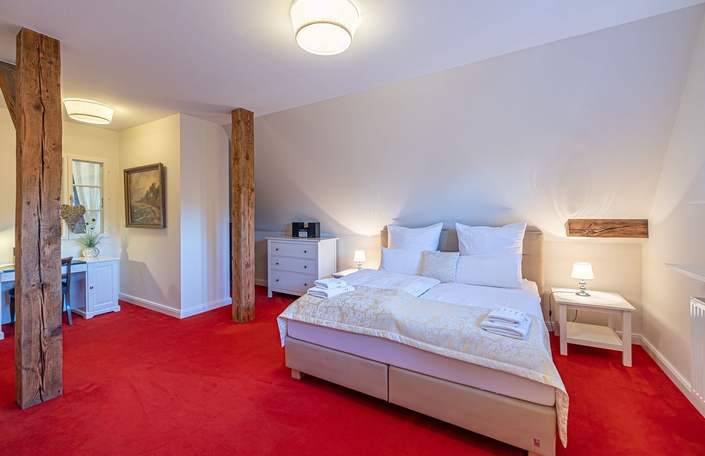 A bedroom with a large bed, a desk, and a chair. There is a window with curtains and a painting on the wall.