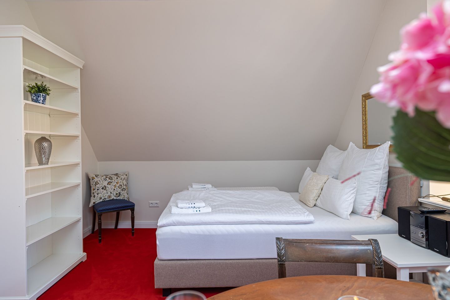 Quarto de hotel com cama, escrivaninha, cadeira, estante e tapete vermelho.