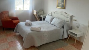 A neatly made bed with pillows and a folded towel, a small bedside table with a lamp, a chair, and a desk with a chair.