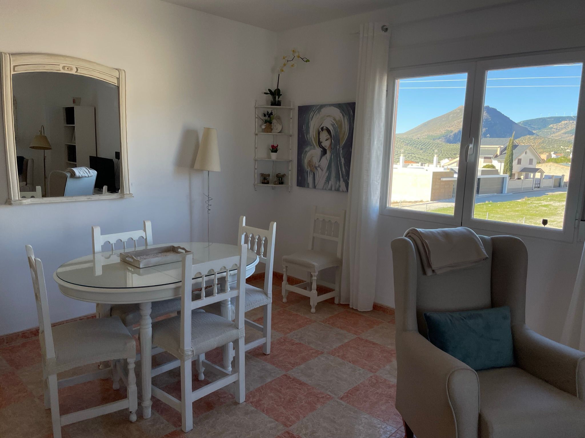 A room with a round glass table, white chairs, a sofa with a blue pillow, a painting, a mirror, and a window with a view of houses and hills.