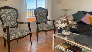 A living room with two patterned armchairs, a glass coffee table, a sofa with cushions, a vase with flowers, and a clock on the wall.