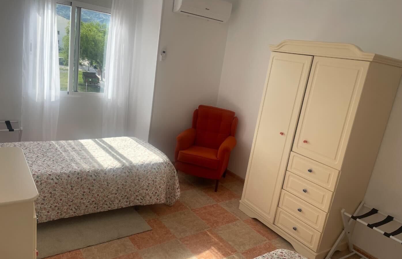 Um quarto com cama, cadeira, guarda-roupa e janela com cortinas.