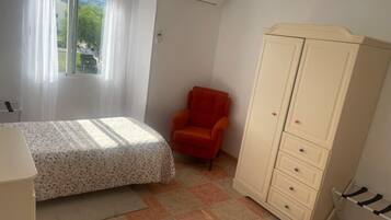Um quarto com cama, cadeira, guarda-roupa e janela com cortinas.