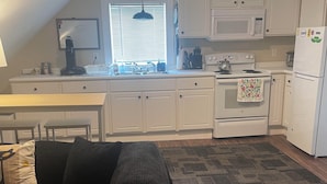 A compact kitchen with white cabinets, a microwave, a stove, and a refrigerator. There's a dining table with chairs, a black sofa, and a patterned rug. - Entire rental unit in Warren, Rhode Island Unit 3 (Warren)