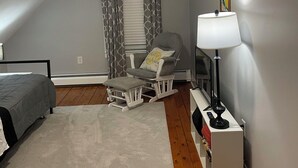 A bedroom with a bed, a rocking chair, a small table with a lamp, a window with curtains, and a picture on the wall. - Entire rental unit in Warren, Rhode Island Unit 3 (Warren)