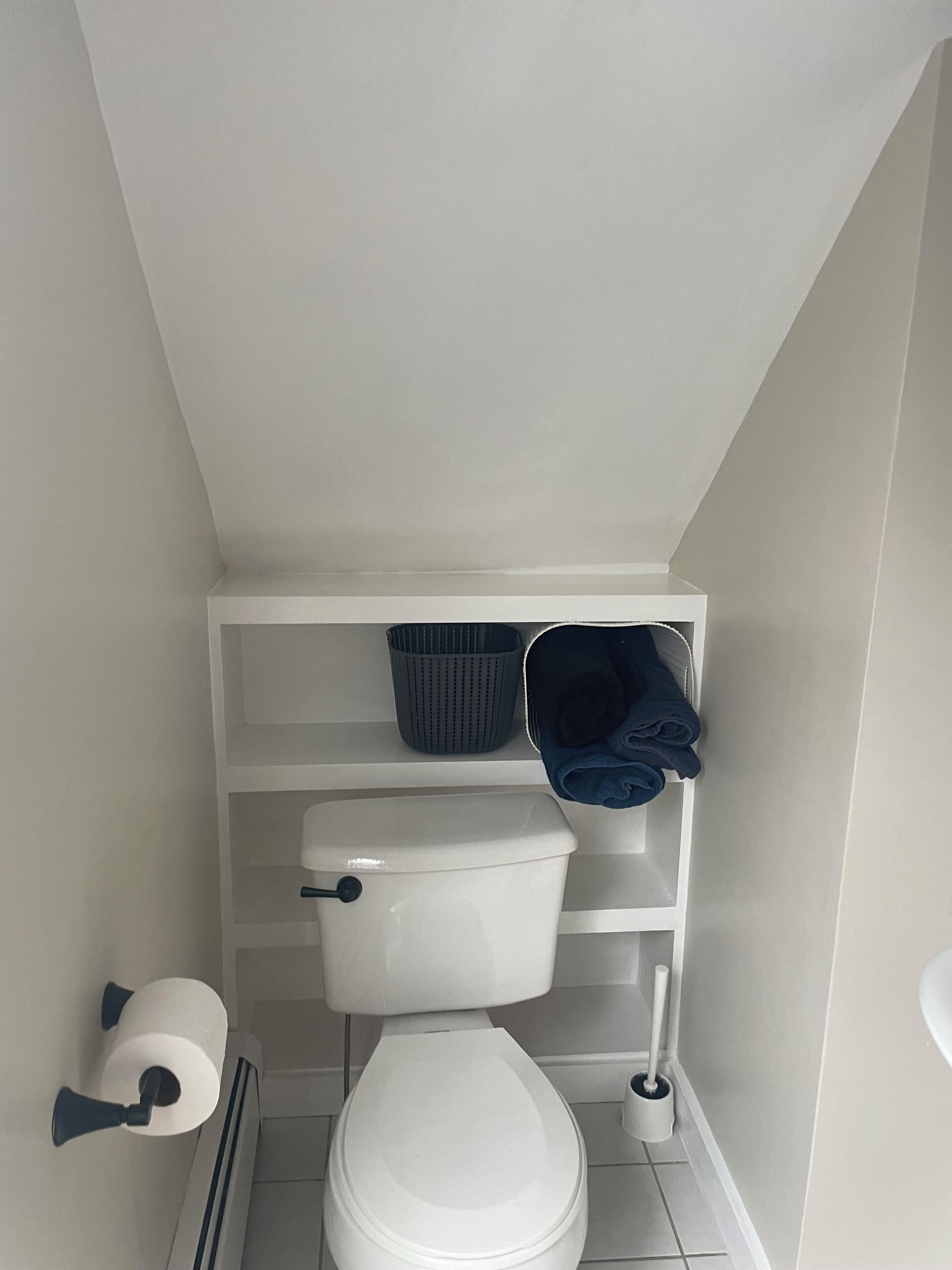 Une petite salle de bain propre, équipée de toilettes, d’une étagère avec un panier et des serviettes, et d’un porte-rouleau de papier toilette.