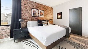A modern bedroom with a brick wall, a large bed, a nightstand, a lamp, and a door.