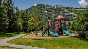 A playground with slides and climbing structures, surrounded by a grassy area and trees, with houses in the background on a hillside. - On Top Of Everything at Beech (Beech Mountain)