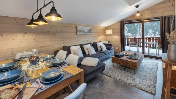Un accogliente soggiorno con parete in legno, un divano, un tavolo da pranzo per quattro persone e un balcone con vista sugli alberi.
