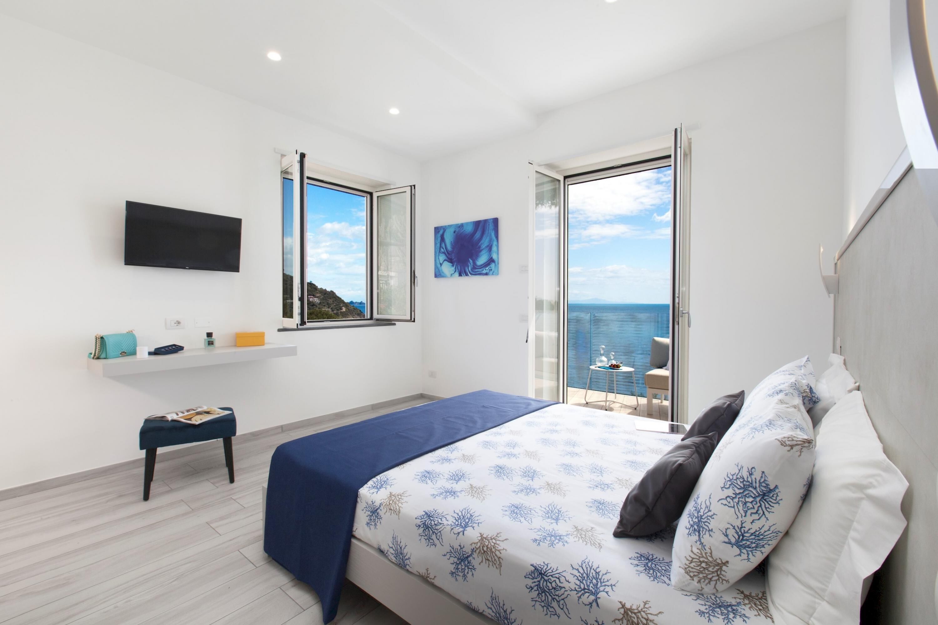 A bedroom with a bed, a TV, a small table, and a window with a view of the sea.