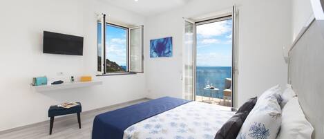 A bedroom with a bed, a TV, a small table, and a window with a view of the sea.
