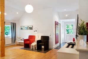 Una habitación moderna con suelo de madera, una silla roja, una silla negra, una mesa blanca y un ventanal con cortinas.