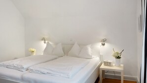 A white double bed with pillows, a bedside table with a radio and vase, and wall-mounted lights.