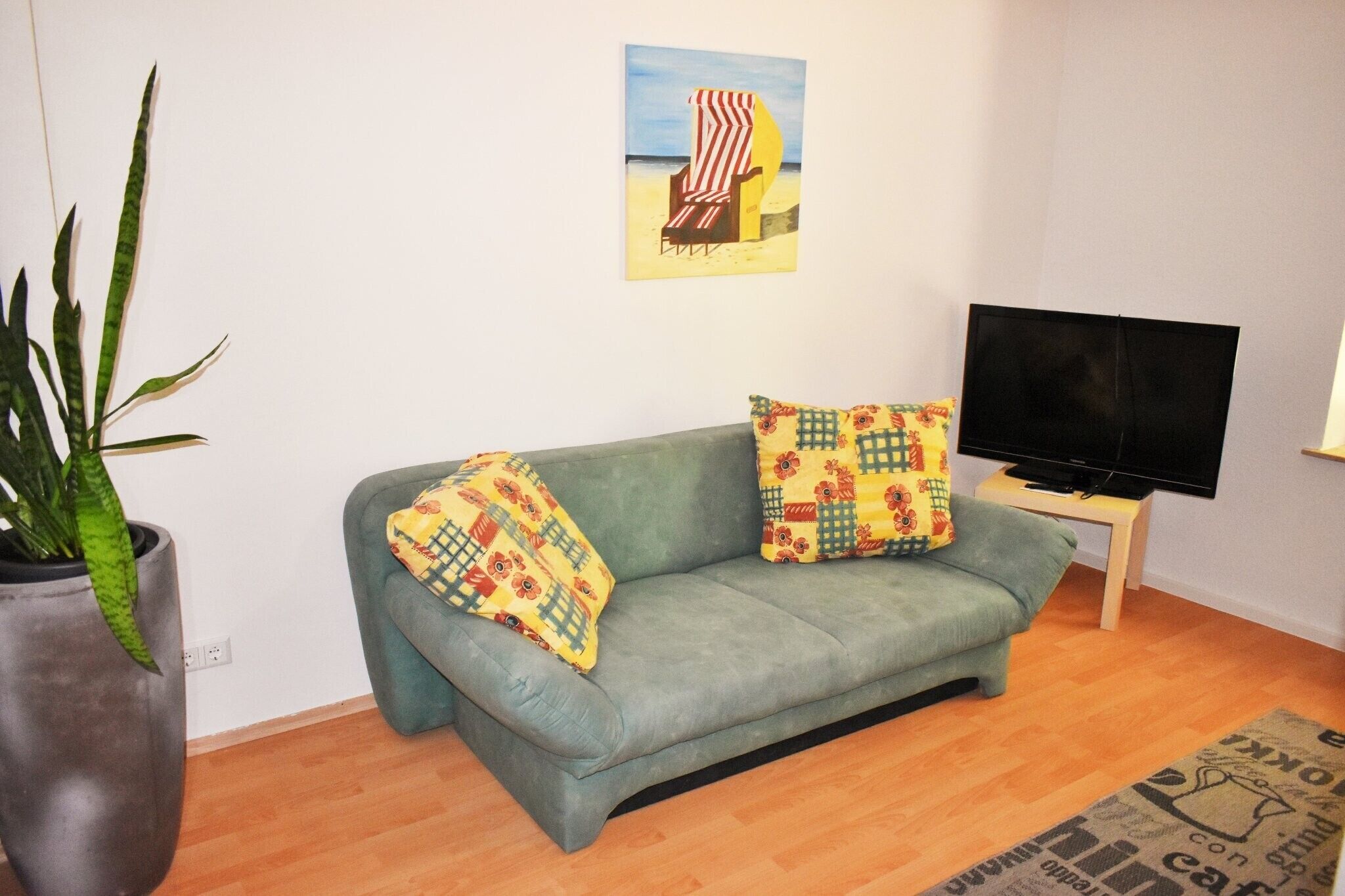 A living room with a sofa, a television, a potted plant, and a beach-themed painting on the wall.