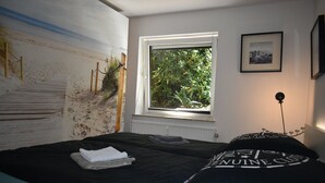 Une chambre à coucher avec un lit, une fenêtre donnant sur un paysage verdoyant, un tableau encadré au mur et un radiateur.