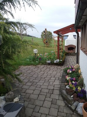 A paved patio with a brick oven, a wooden pergola, and a garden with flowers and a bird feeder.