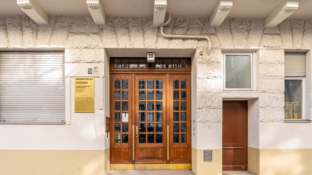 Une entrée d’immeuble avec une porte double en bois, une plaque numérotée au-dessus de la porte et un volet fermé sur le côté gauche.