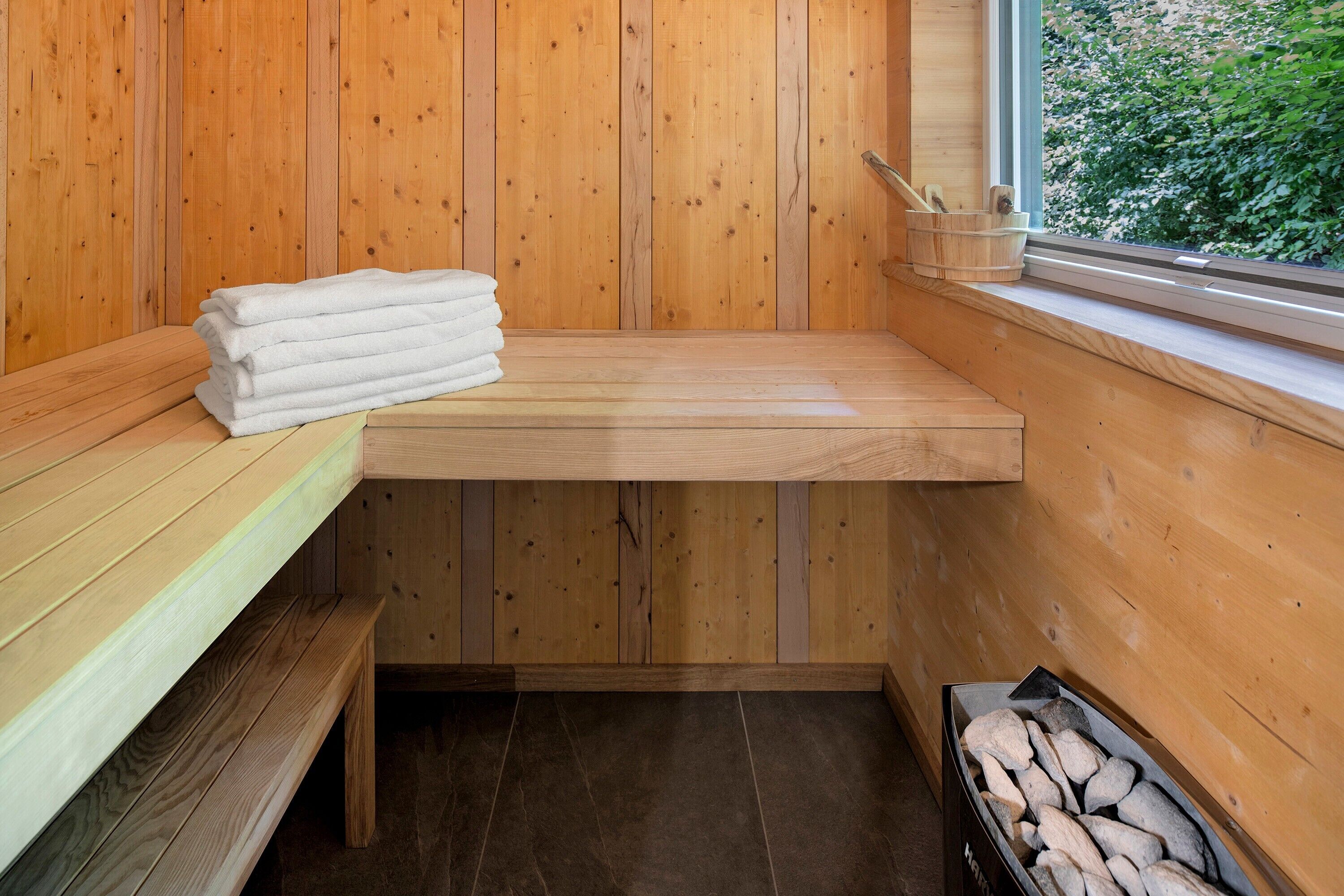 A wooden sauna with benches, a shelf with towels, a window with a view of greenery, and a container with stones.