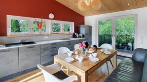 A modern kitchen with a wooden ceiling, a dining area with a table set for four, and large windows offering a view of greenery.