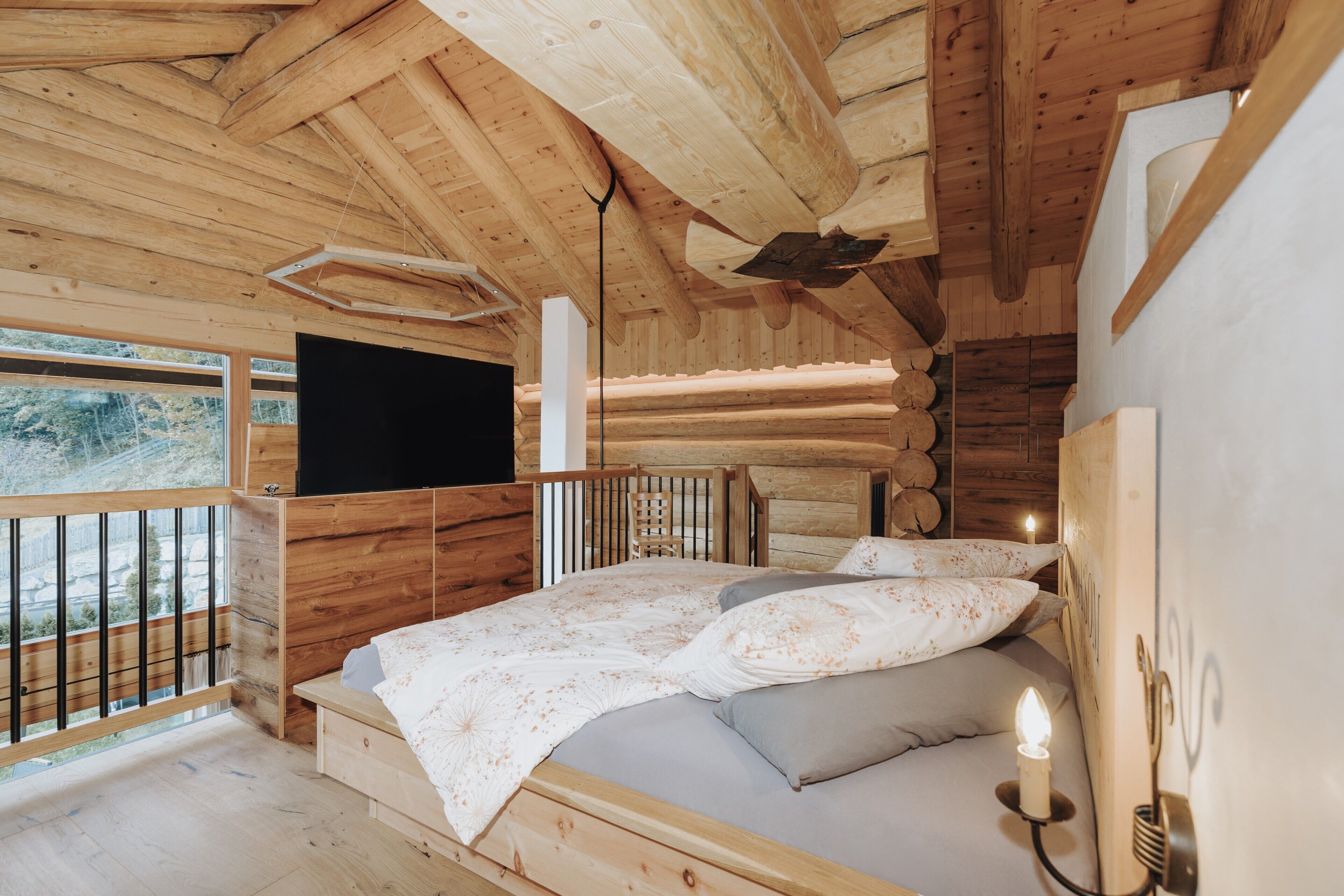 A wooden cabin bedroom with a bed, a TV, and a window overlooking a forest.