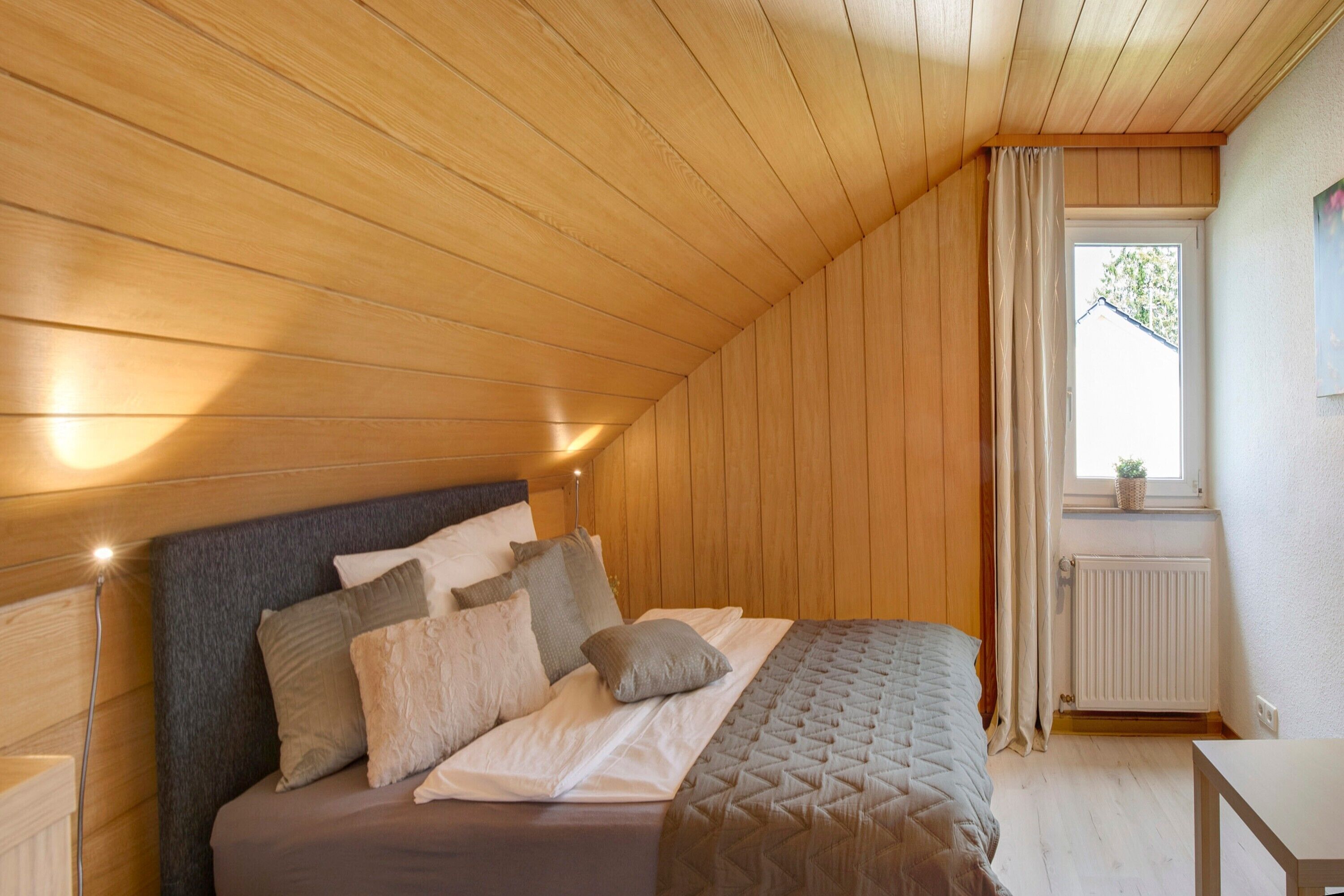A bedroom with a wooden ceiling, a bed with multiple pillows, a bedside table, and a window with curtains.