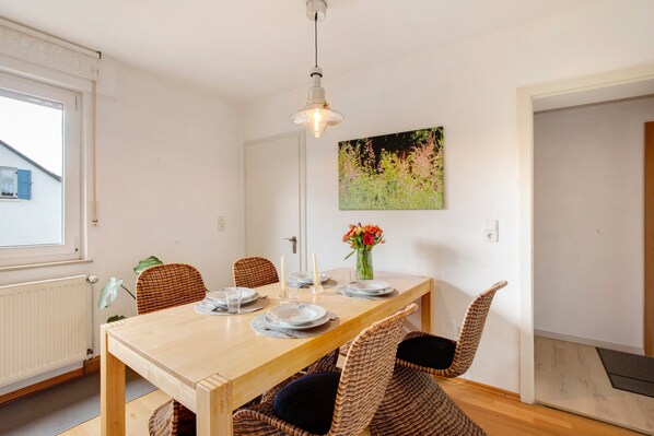 A dining area with a wooden table, wicker chairs, a pendant light, and a floral painting on the wall.