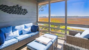 A covered outdoor seating area with a wicker sofa and ottoman, a white tray table, and a view of a field through large windows.