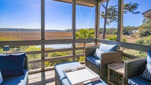 A covered outdoor seating area with wicker furniture and cushions, overlooking a body of water and a distant landscape.