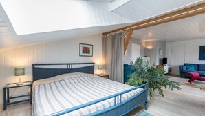 Une chambre avec une lucarne, un lit recouvert d’une couvre-lit à rayures, des lampes de chevet, une plante en pot et une vue sur le salon.