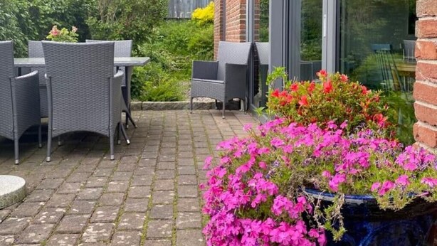 Une terrasse extérieure avec un sol en briques, une table entourée de chaises et une grande fenêtre donnant sur un jardin.