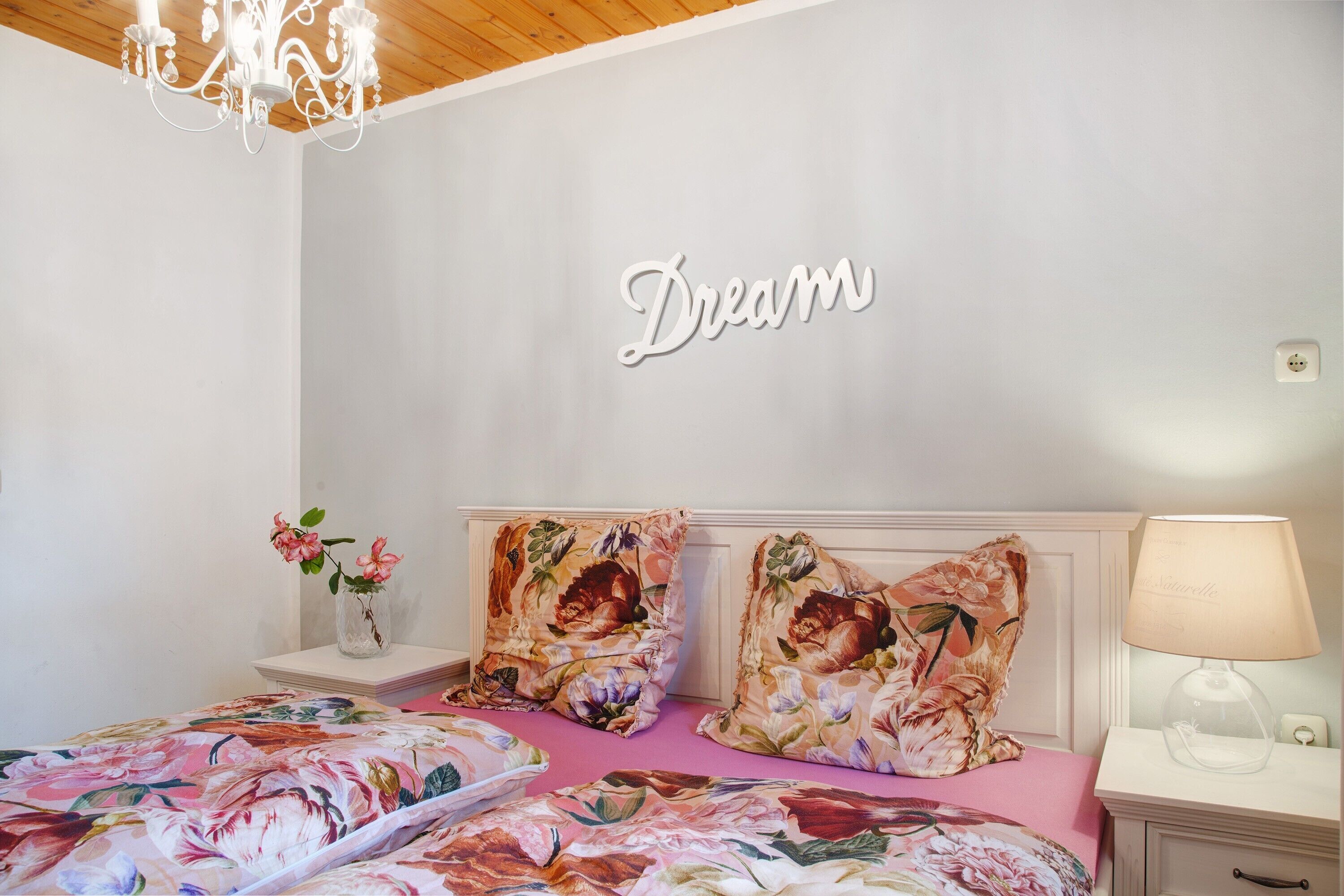 A bedroom with a floral bedspread, two matching pillows, a bedside table with a lamp, and a wall-mounted word 'Dream'.