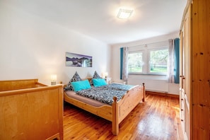 Una cama de madera con cabecera, una mesita de noche con lámpara, una ventana con cortinas e imagen colgada en la pared.