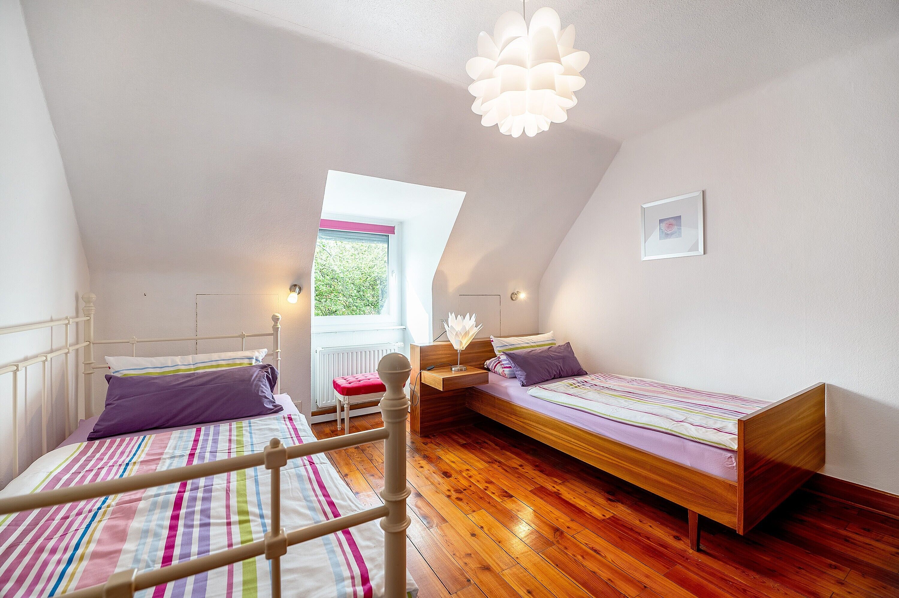 A bedroom with two beds, a window, a wooden floor, and a ceiling light fixture.