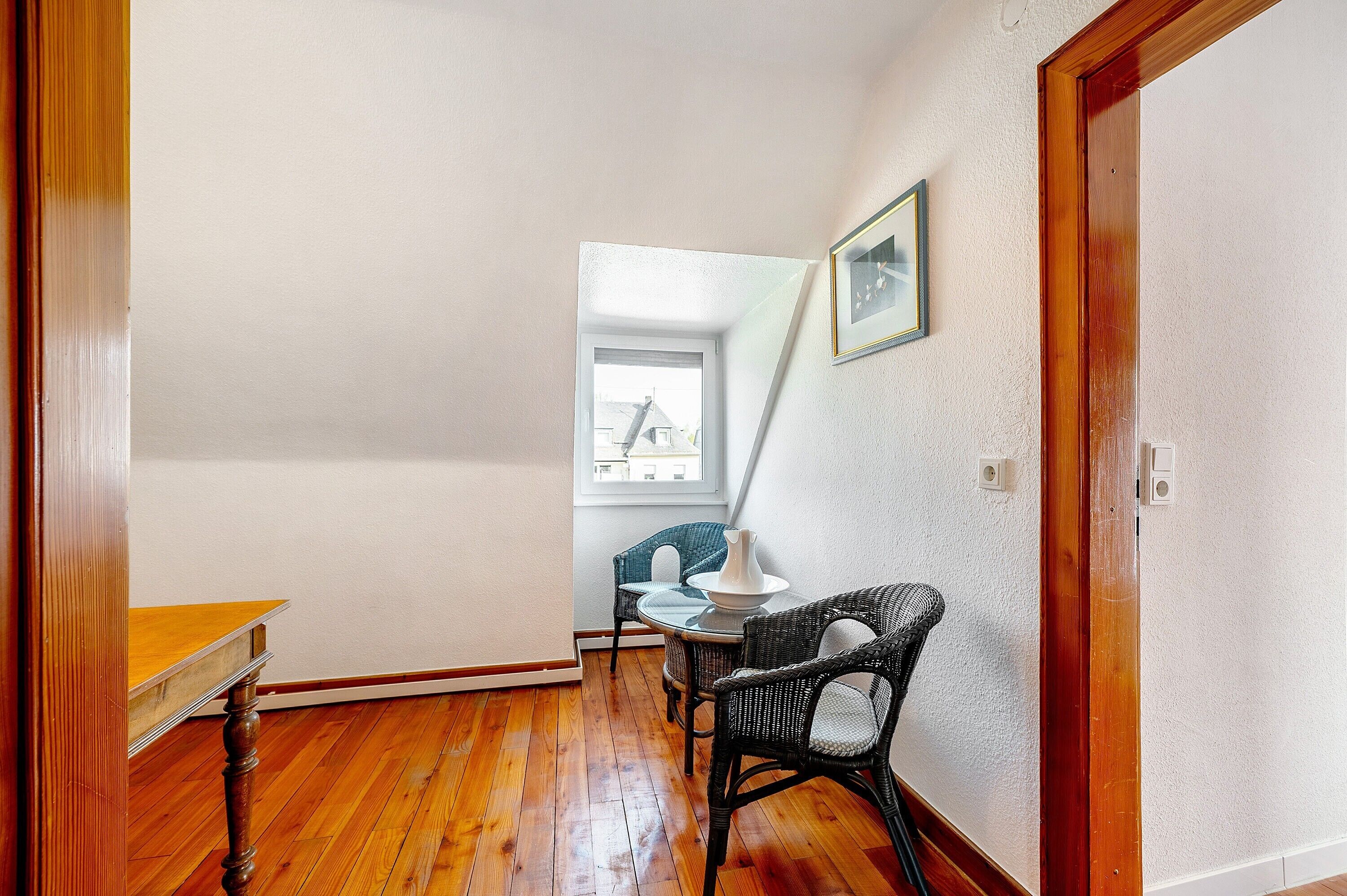 A room with a wooden floor, a table, two wicker chairs, a white lamp, and a framed picture on the wall.