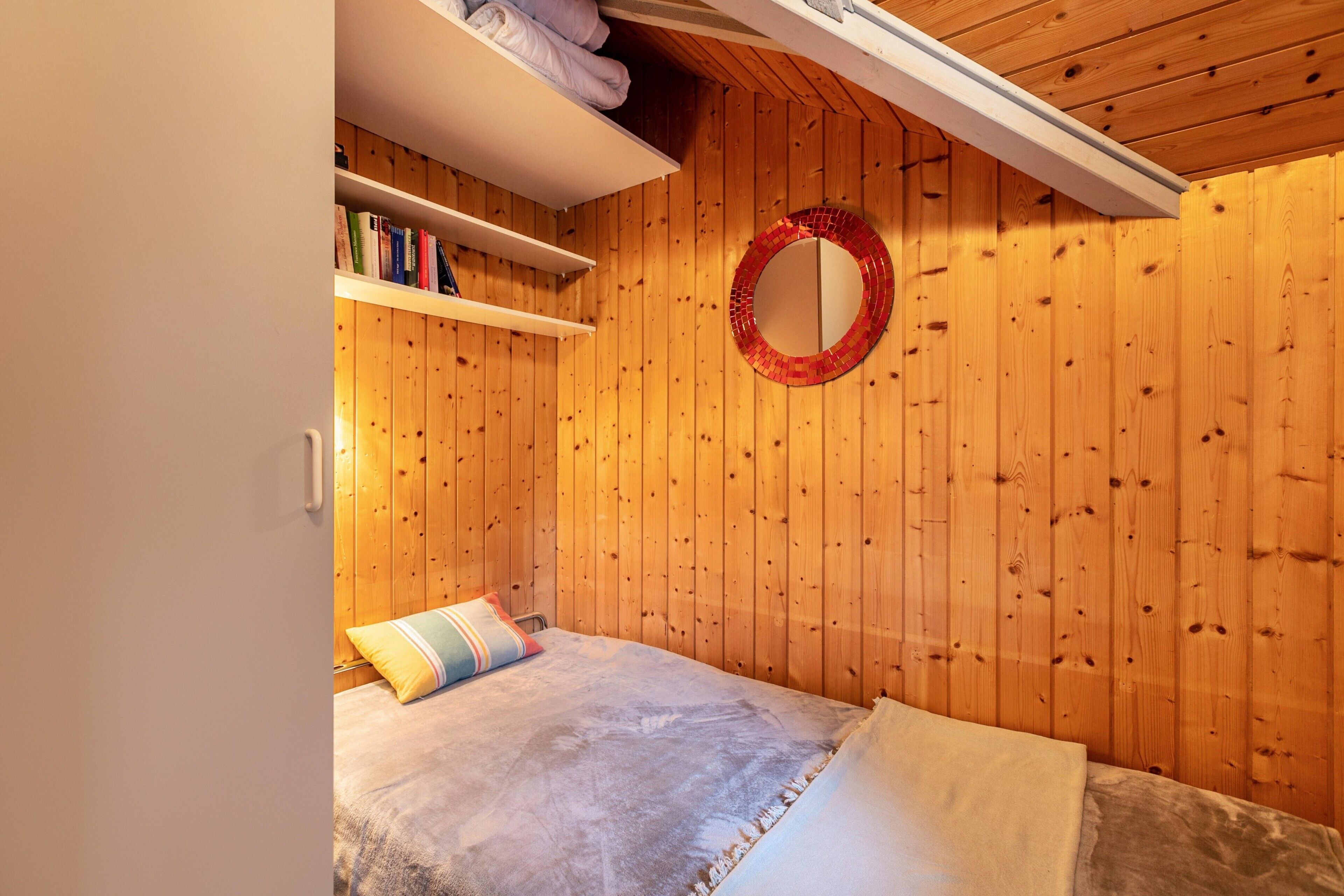 A wooden-paneled room with a bed, a shelf with books, and a circular mirror on the wall.