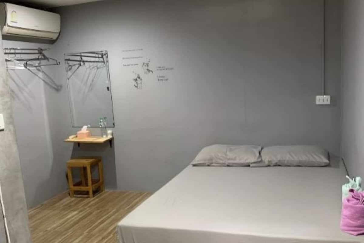 A bedroom with a bed, a wooden floor, a small table with a chair, a wall-mounted air conditioner, and a towel rack.