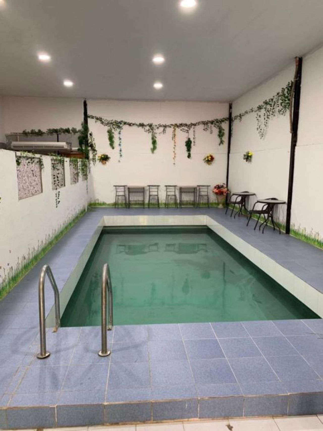 Une piscine dotée d’une terrasse carrelée, entourée de chaises et de tables, et agrémentée de plantes et de fleurs décoratives sur les murs.