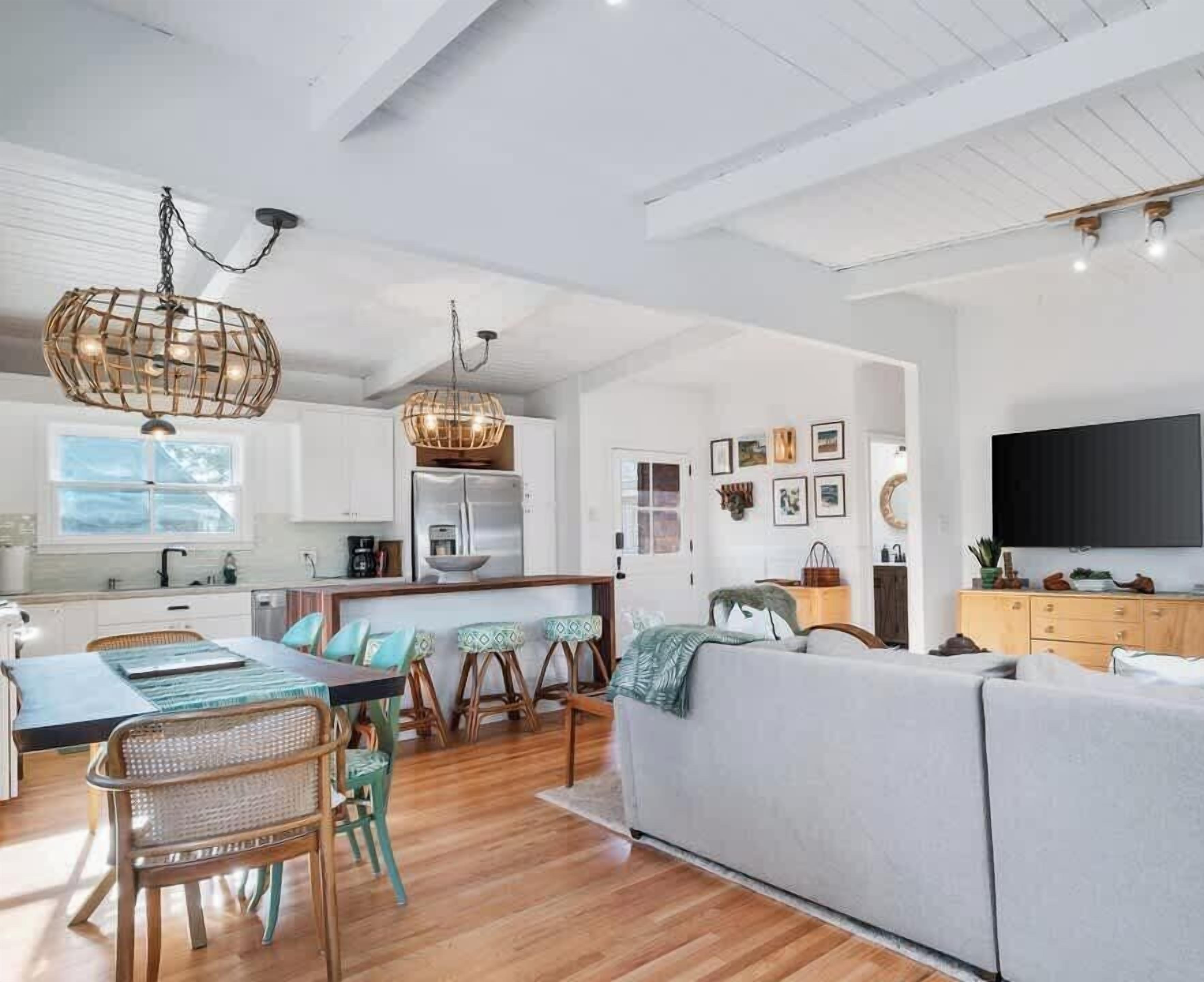 A spacious living area with a dining table, chairs, and a sofa. There is a kitchen area with a refrigerator and a hanging light fixture.