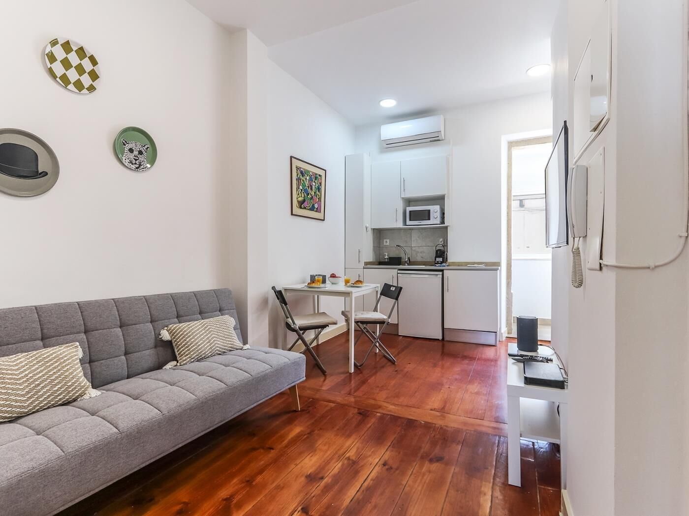 Un espacio habitable compacto con un sofá gris, una pequeña mesa de comedor y una kitchenette.