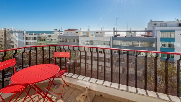 A balcony with a red table and chairs, overlooking a cityscape with modern buildings and a clear sky.