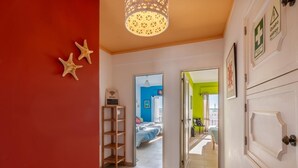 A hallway with a red wall, a wooden shelf with a WiFi sign, and a door leading to a room with a bed and a blue wall.