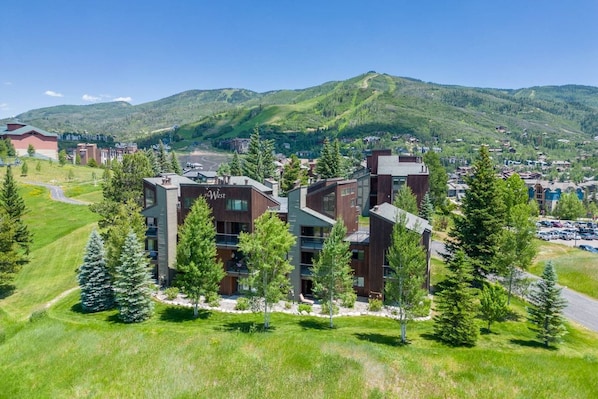 A modern multi-story building with a flat roof, surrounded by green trees and a hilly landscape in the background.