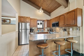 A kitchen with wooden cabinets, a stainless steel refrigerator, a granite countertop, and bar stools.