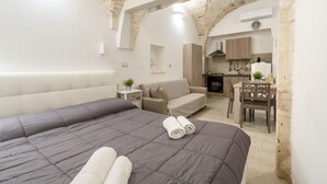 A bedroom with a large bed, a sofa, a small table, and a kitchen area visible through an archway. - GuestHost - Casa Vita 49 in Ceglie Messapica (Ceglie Messapica)