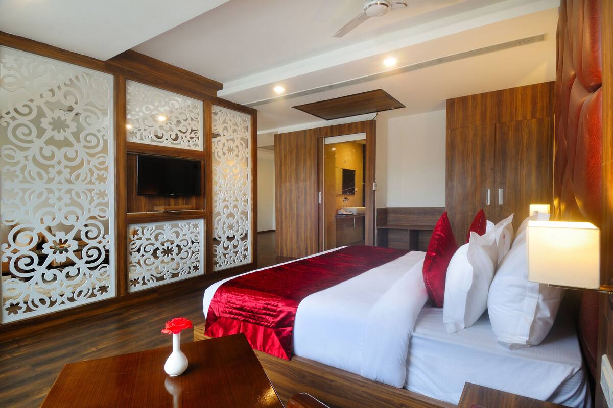 Una habitación de hotel con suelo de madera, una cama con ropa de cama roja y blanca, un televisor montado en la pared y un panel decorativo blanco.