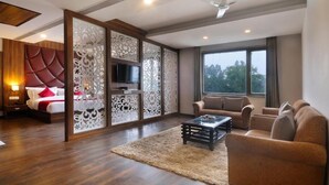 Quarto de hotel moderno com uma cama grande, televisão, conjunto de sofás e uma janela ampla.