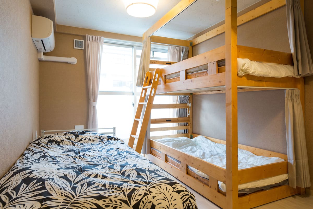Une chambre avec un lit superposé, une échelle, une fenêtre avec des rideaux et un lit recouvert d’une couvre-lit à motifs floraux.