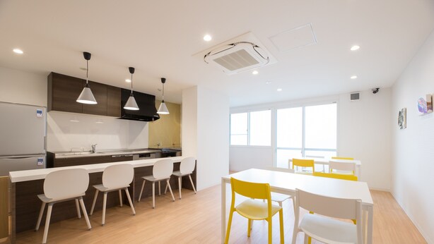 Une cuisine moderne avec des armoires blanches, une hotte noire et des luminaires suspendus. On y trouve également un coin repas avec une table blanche et des chaises jaunes.