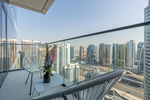 A balcony with a white chair and a vase of flowers, overlooking a cityscape with modern high-rise buildings.